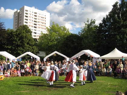 Foto der Festwiese auf dem Dorffest, auf dieser findet ein Tanzauftritt statt. Die Frauen tragen weiß-rote Trachten. Im Hintergrund ist ein Hochhaus zu sehen.