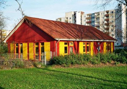 Ein einstöckiges Haus mit einer strahlend-farbigen Außenfassade aus roter Holzvertäfelung und gelben Fensterrahmen. Die Fenster sind mit ebenfalls mit gelben Fensterläden aus Holz bestückt.