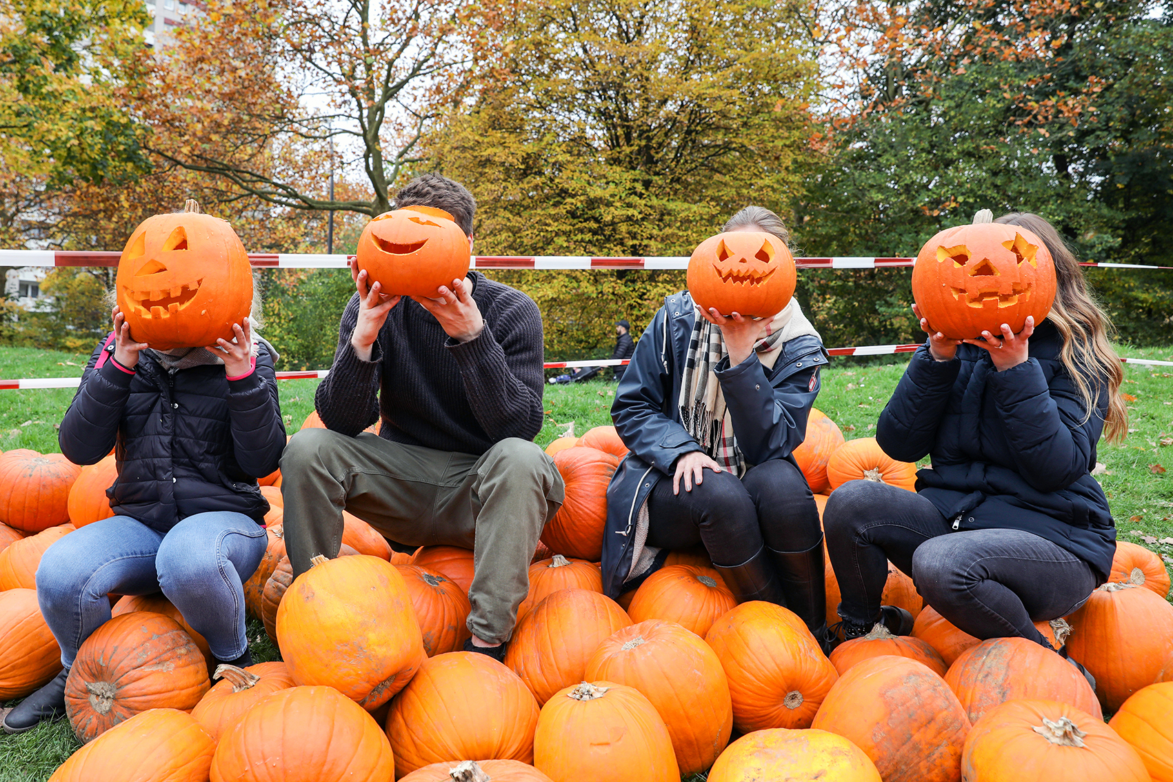 Auf einer grünen Wiese liegt ein großer Haufen orangener Kürbisse. Auf diesen Kürbissen sitzen vier Personen in einer Reihe. Jede Person hält sich einen geschnitzten Halloween-Kürbis vor das Gesicht. Alle Kürbisse lachen freundlich.