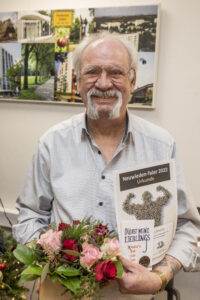 Foto eines Preisträgers des "Neuwieden - Taler 2022". In der linken Hand hält er den Blumenstrauß, in der rechten Hand zeigt er die Urkunde und den Taler in die Kamera. Der Mann trägt eine Brille und grauen Schnurrbart.