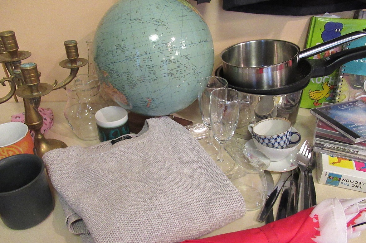 Das Foto zeigt einen Tisch mit diversen Objekten für einen Flohmarkt. U.a. einen Kronleuchter, einen Globus, Geschirr und Besteck sowie CD's und Bücher.