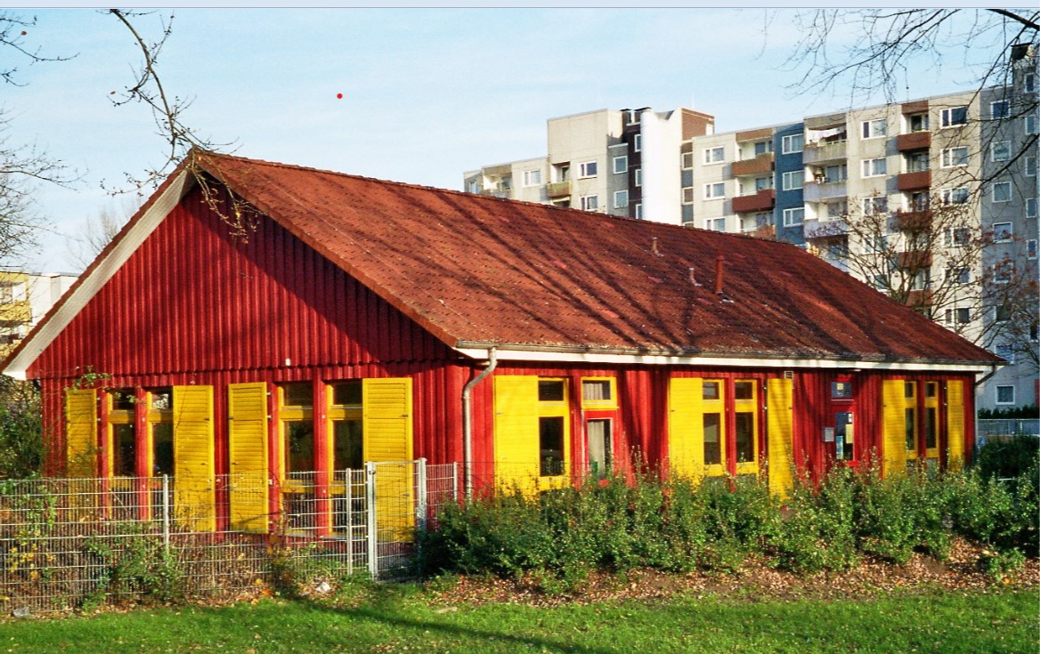 Ein einstöckiges Haus mit einer strahlend-farbigen Außenfassade aus roter Holzvertäfelung und gelben Fensterrahmen. Die Fenster sind mit ebenfalls mit gelben Fensterläden aus Holz bestückt.