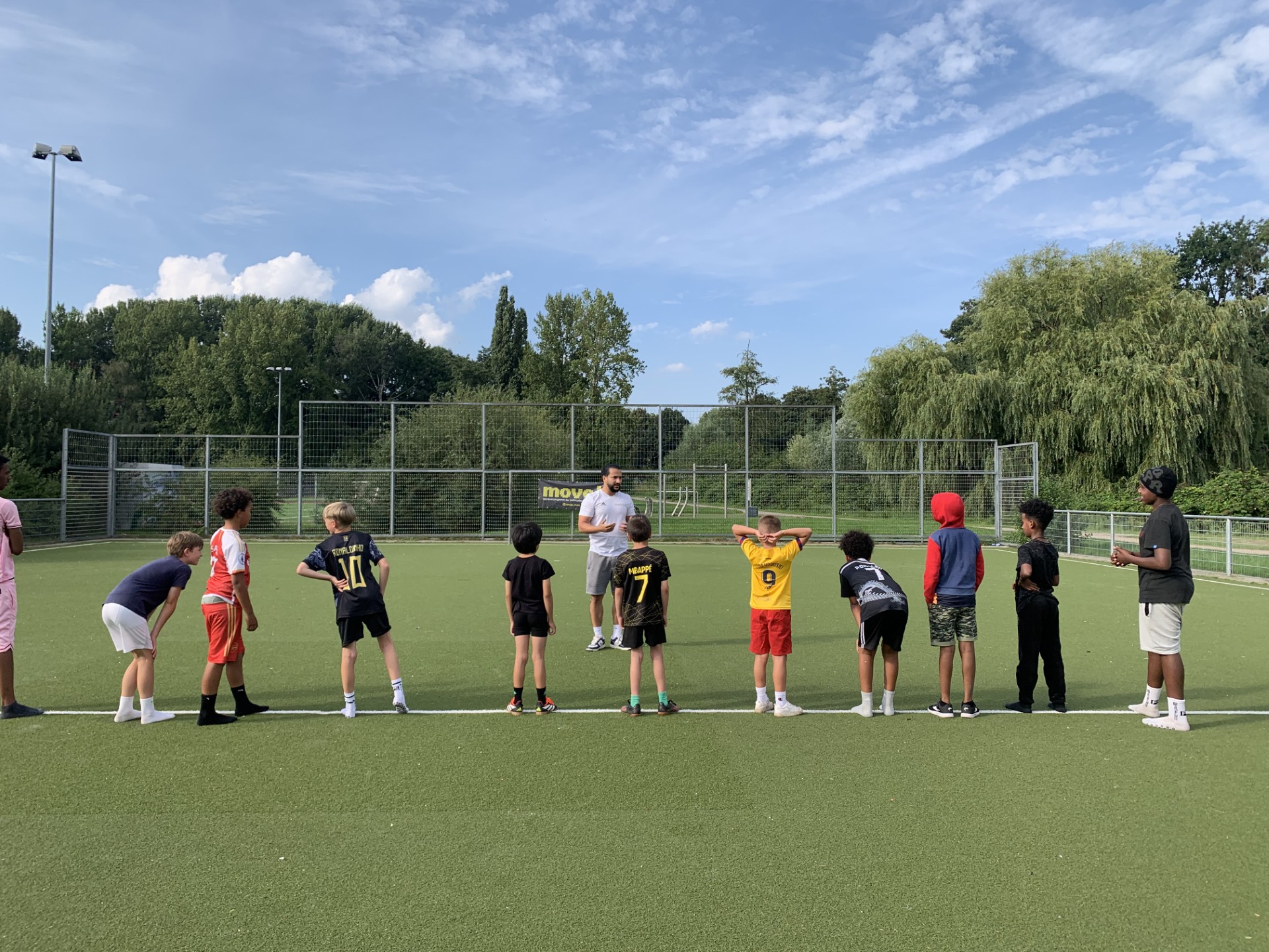 Bild zeigt einen Sportplatz, auf dem ein Trainer zu verschiedenen Kindern spricht, die Fußball spielen wollen