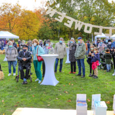 Auf einer Festwiese stehen weiße Pavillons und weiße Stehtische. Die Menschen stehen vor einem Pavillon und blicken in eine Richtung auf eine Leinwand.