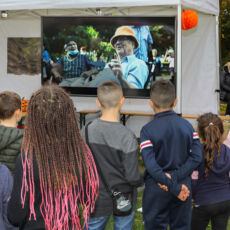Kinder stehen mit dem Rücken zur Kamera vor einem weißen Pavillon. Im Pavillon wird auf einer Leinwand ein Film gezeigt.