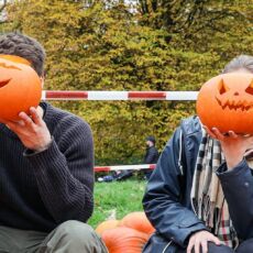 Auf einer grünen Wiese liegt ein großer Haufen orangener Kürbisse. Auf diesen Kürbissen sitzen vier Personen in einer Reihe. Jede Person hält sich einen geschnitzten Halloween-Kürbis vor das Gesicht. Alle Kürbisse lachen freundlich.