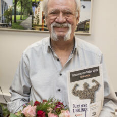 Foto eines Preisträgers des "Neuwieden - Taler 2022". In der linken Hand hält er den Blumenstrauß, in der rechten Hand zeigt er die Urkunde und den Taler in die Kamera. Der Mann trägt eine Brille und grauen Schnurrbart.