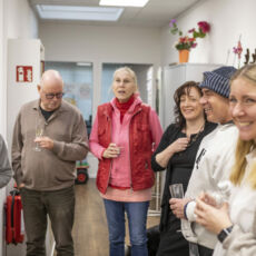 Sechs Personen stehen in einem Halbkreis mit Sektgläsern in der Hand und gucken lächelnd in die gleiche Richtung, zum linken Bildrand am Betrachter des Fotos vorbei.