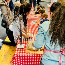 An einem langen Tisch sitzen mehrere Kinder vor Teigrollern und Keksausstechern.