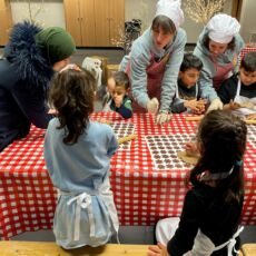 An einem langen Tisch sitzen mehrere Kinder vor Teigrollern und Keksausstechern. Zwei Erwachsene Personen mit rotkarierter Schürze und weißer Kochmütze zeigen ihnen, wie man den Keksteig ausrollt.