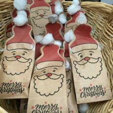 In einem geflechteten Korb liegen kleine braune Papierbeutel mit einem Weihnachtsmann-Aufdruck und dem Schriftzug "Merry Christmas". Der Beutel hat oben die Form einer Zipfelmütze, diese ist rot abgedruckt und der weiße Bommel ist aus Watte aufgeklebt.