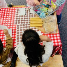 Das Foto zeigt einen Tisch mit rot-weiß karierter Wachsdecke von oben. An diesem Tisch sitzen Kinder und verzieren Kekse. Oben links wir ihnen ein silbernes Tablett angereicht. Auf diesem Tablett sind kleine Gläser mit bunten Perlen aufgereiht, aus denen die Kinder mit kleinen Löffeln schöpfen können.