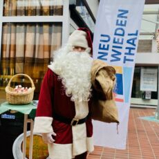 Im überdachten Bereich des EKZ Rehrstieg-Galleria posiert ein Mann. Dieser ist verkleidet als Nikolaus, mit weißem Bart und Jutesack über der Schulter und steht vor einer Beachflag. Diese trägt die Aufschrift "Neuwiedenthal".