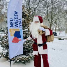 Ein verkleideter Nikolaus steht draußen im Schnee, vor einer beleuchteten Tanne.